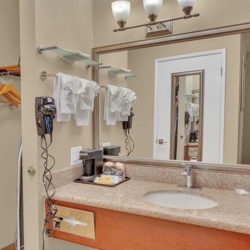 Bathroom vanity area with a sink, mirror, hair dryer, towels, and toiletries alongside a small wardrobe and an ironing board.