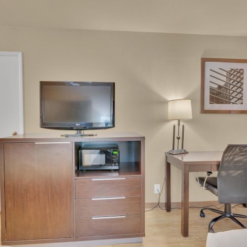 The image shows a hotel room with a TV on a dresser, a desk with a chair, and a lamp, next to a framed picture on the wall.