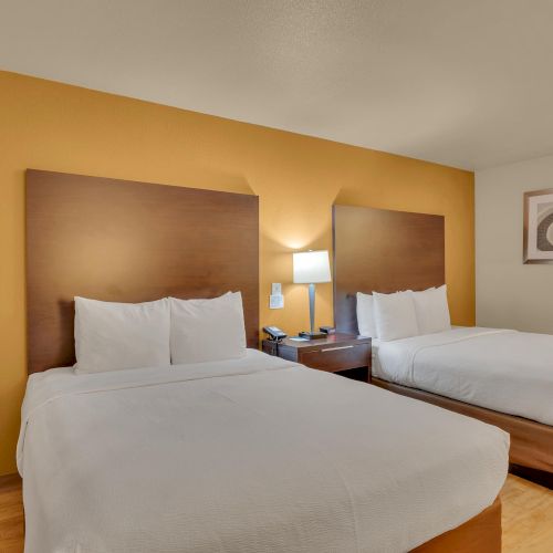 The image shows a hotel room with two beds, a bedside table with a lamp, and wall art in a neutral and cozy setting.