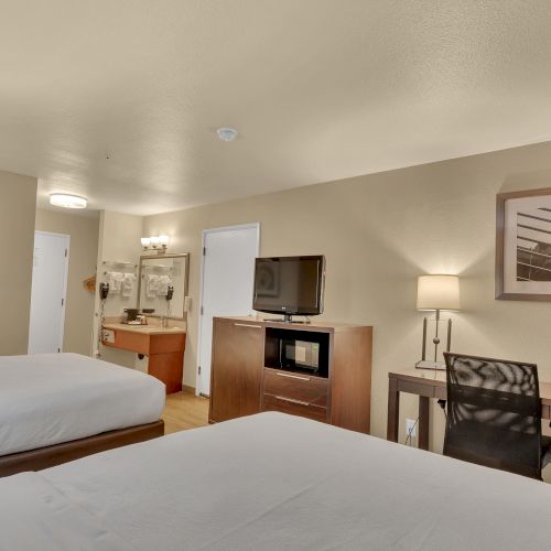 The image shows a hotel room with two beds, a TV on a dresser, a desk with a chair, and framed artwork on the walls.