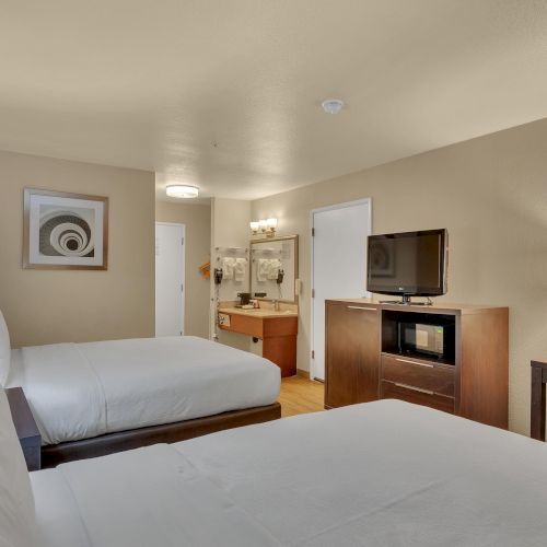 The image shows a hotel room with two beds, a TV, a microwave, a desk, and a lamp, creating a cozy and functional space.