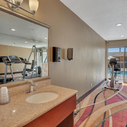 The image shows a gym with exercise equipment, a water cooler, and a mirror, adjacent to a pool area visible through a sliding door.