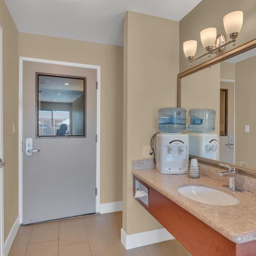 The image shows a bathroom with a sink, mirror, soap dispenser, and a water cooler on the counter, and a door in the background.
