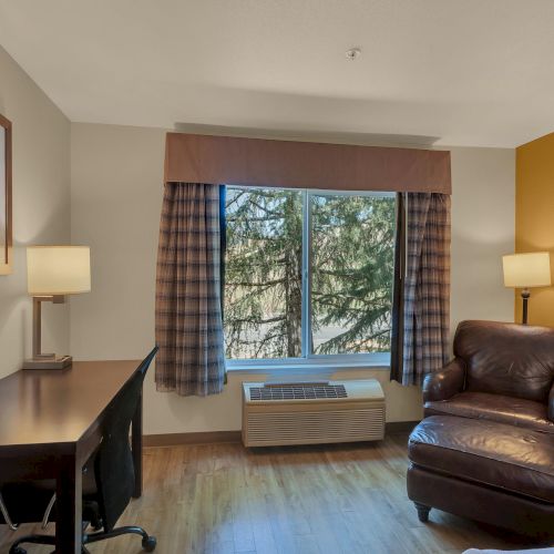The image shows a hotel room with a desk, chair, and lamp on one side, an armchair on the other, and a window with a nature view.