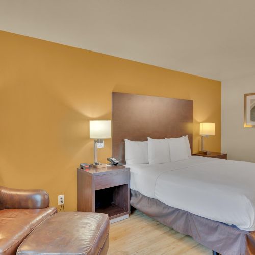 A hotel room with a double bed, two lamps, a large armchair, and artwork on a mustard-colored accent wall, creating a cozy atmosphere.
