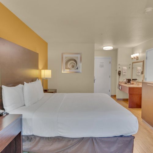 A hotel room with a king-sized bed, nightstands, lamps, TV, mini fridge, and a view into the bathroom area, featuring neutral decor.