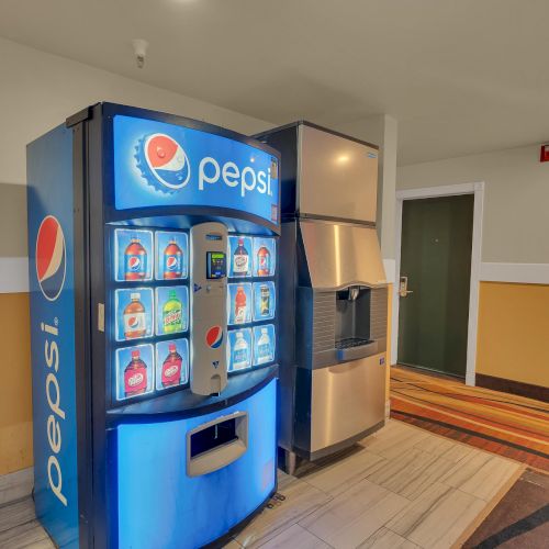 A Pepsi vending machine and an ice dispenser are in a hallway with beige walls and a mix of carpet and tile flooring.