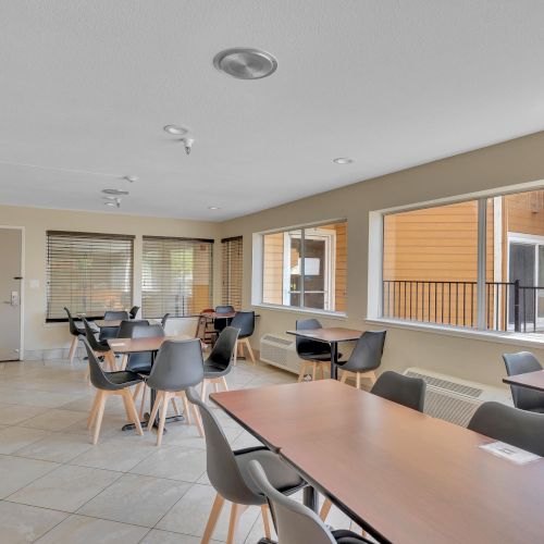 The image shows an empty dining area with tables, chairs, and large windows overlooking the outside of a building.
