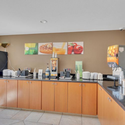 A hotel breakfast area with dispensers, coffee machines, cups, and food items on a counter against a beige wall displaying fruit artwork.