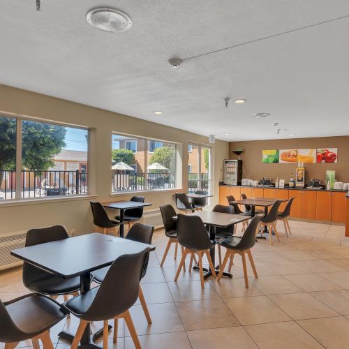 The image shows a dining area with tables and chairs, large windows, and a counter with food and drink options, likely a breakfast area.