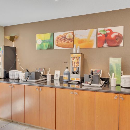 The image shows a breakfast buffet area with coffee machines, juice dispenser, stack of bowls, and wall art of food and fruit decorations.