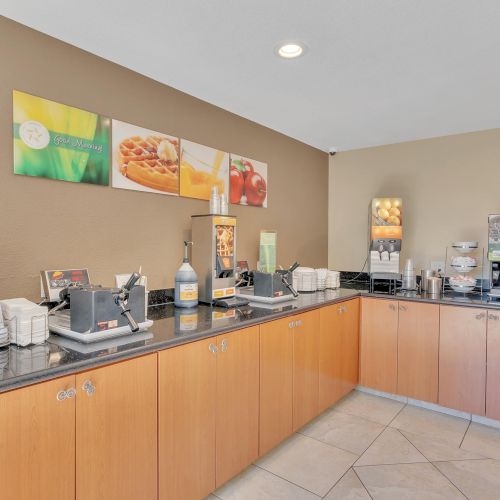 The image shows a modern breakfast buffet setup with various dispensers, plates, cups, and food items on a counter in a dining area.