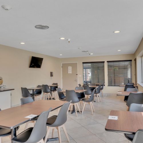 The image shows a dining area with tables, chairs, a countertop with dispensers, and a wall-mounted TV. Large windows provide natural light.