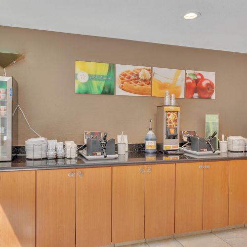 The image shows a breakfast buffet setup with a juice machine, coffee dispensers, cups, and condiments, against a wall with food-themed art.