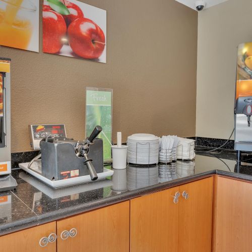 The image shows a breakfast buffet with a waffle maker, juice machine, and stack of plates and cups on a counter.