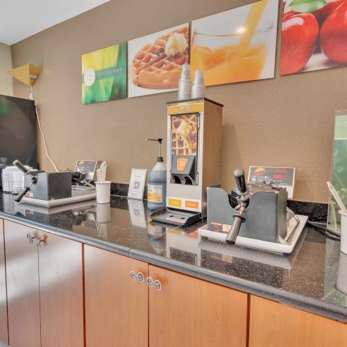 A breakfast buffet setup with a waffle maker, juice dispenser, and various utensils on a countertop, featuring fruit images on the wall.