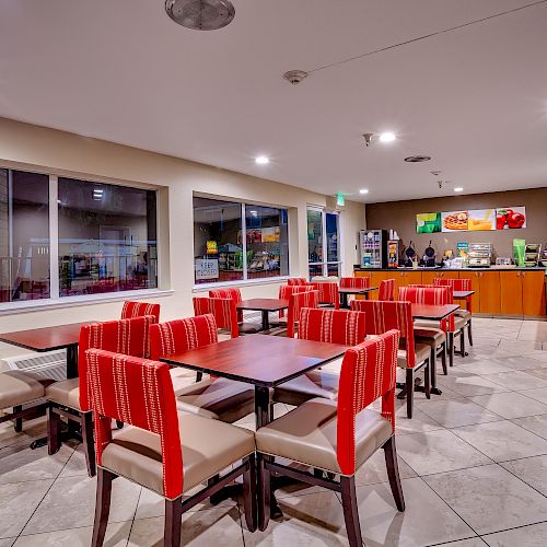 The image shows a dining area with red and beige chairs, multiple tables, large windows, and a counter with various food and drink items.