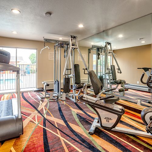 The image shows a gym with a treadmill, a multi-function exercise machine, an exercise bike, and another cardio machine on a colorful carpet.