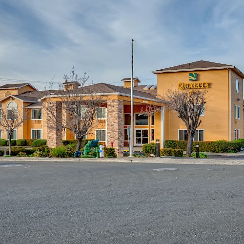 A hotel building with a yellow and beige exterior, surrounded by trees, a clear sky, and an empty parking lot.