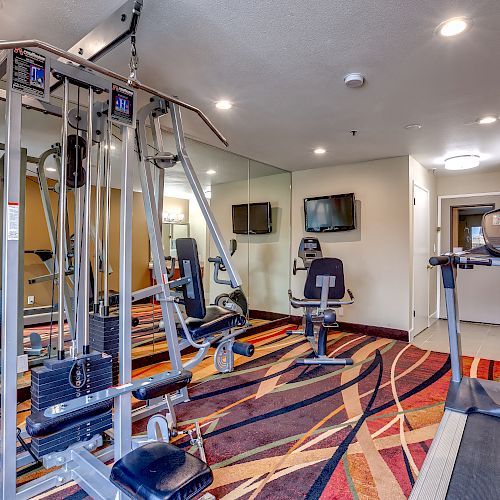 The image shows a small gym room with various exercise equipment, including a cable machine, exercise bike, treadmill, and TV screens.