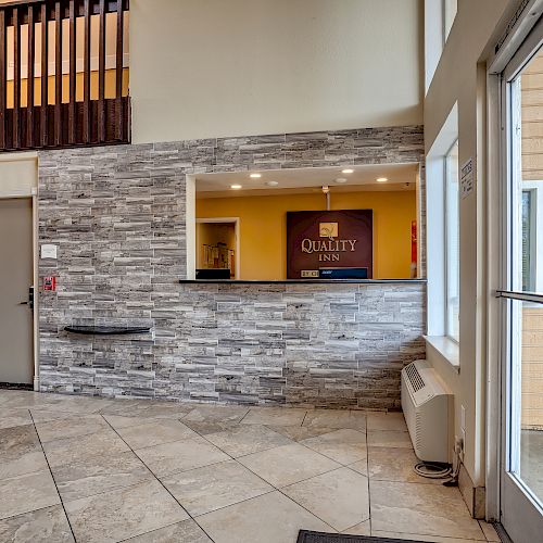 The image shows a hotel lobby with a tiled floor, modern front desk with a "Quality Inn" sign behind, and a large windowed entrance.