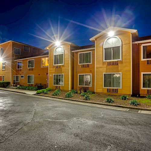 A nighttime view of a modern, well-lit apartment complex with parking spaces, greenery, and accessible parking spots.