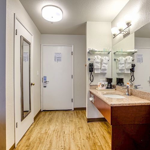 This image shows a hotel bathroom with a sink, large mirror, towels, a coffee maker, and a hairdryer, all under bright and organized lighting.