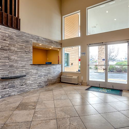 A clean, modern lobby with a stone accent wall, front desk, tiled floor, and large windows letting in natural light, leading to the entrance.