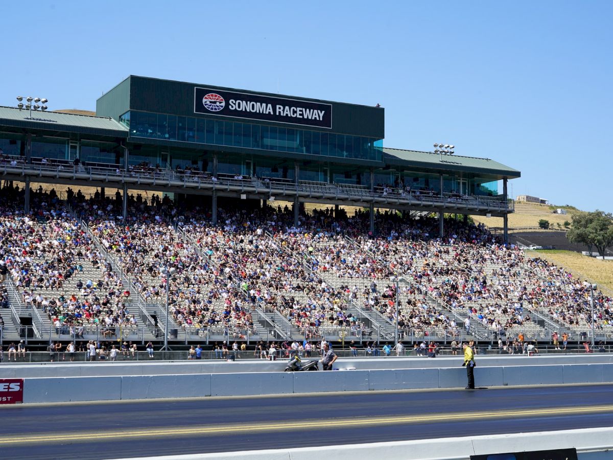 Sonoma Raceway grandstand packed with spectators, overlooking a drag strip on a sunny day, with a clear blue sky in the background.