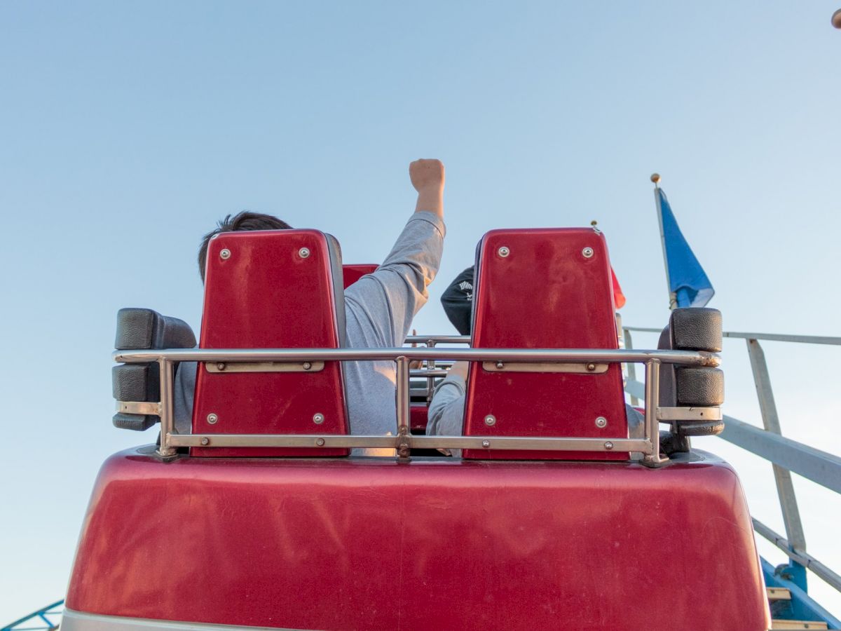 People on a red amusement ride with one person raising their arm, viewed from behind, under a clear blue sky with some flags in the background.