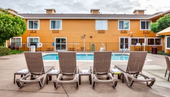 A pool area with four lounge chairs, a fence, and a two-story tan building with several windows and trees on both sides.