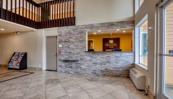 The image shows a hotel lobby with tiled flooring, a front desk with a "Quality Inn" sign, a vending machine, and a staircase leading to the upper level.