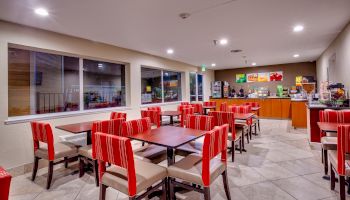 The image shows a well-lit dining area with red and white chairs, wooden tables, a serving counter, and various food and drink dispensers.