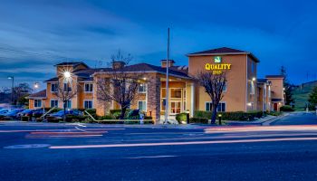 This image shows a Quality Inn hotel building at dusk with a lit sign, parking lot, and streaks of light from passing vehicles in the foreground.