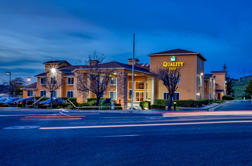 This image shows a Quality Inn hotel building at dusk with a lit sign, parking lot, and streaks of light from passing vehicles in the foreground.