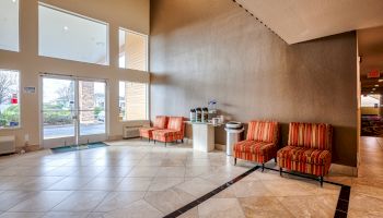 The image shows a spacious lobby with large windows, an entrance door, striped chairs, a coffee station, and tiled flooring, ending the sentence.