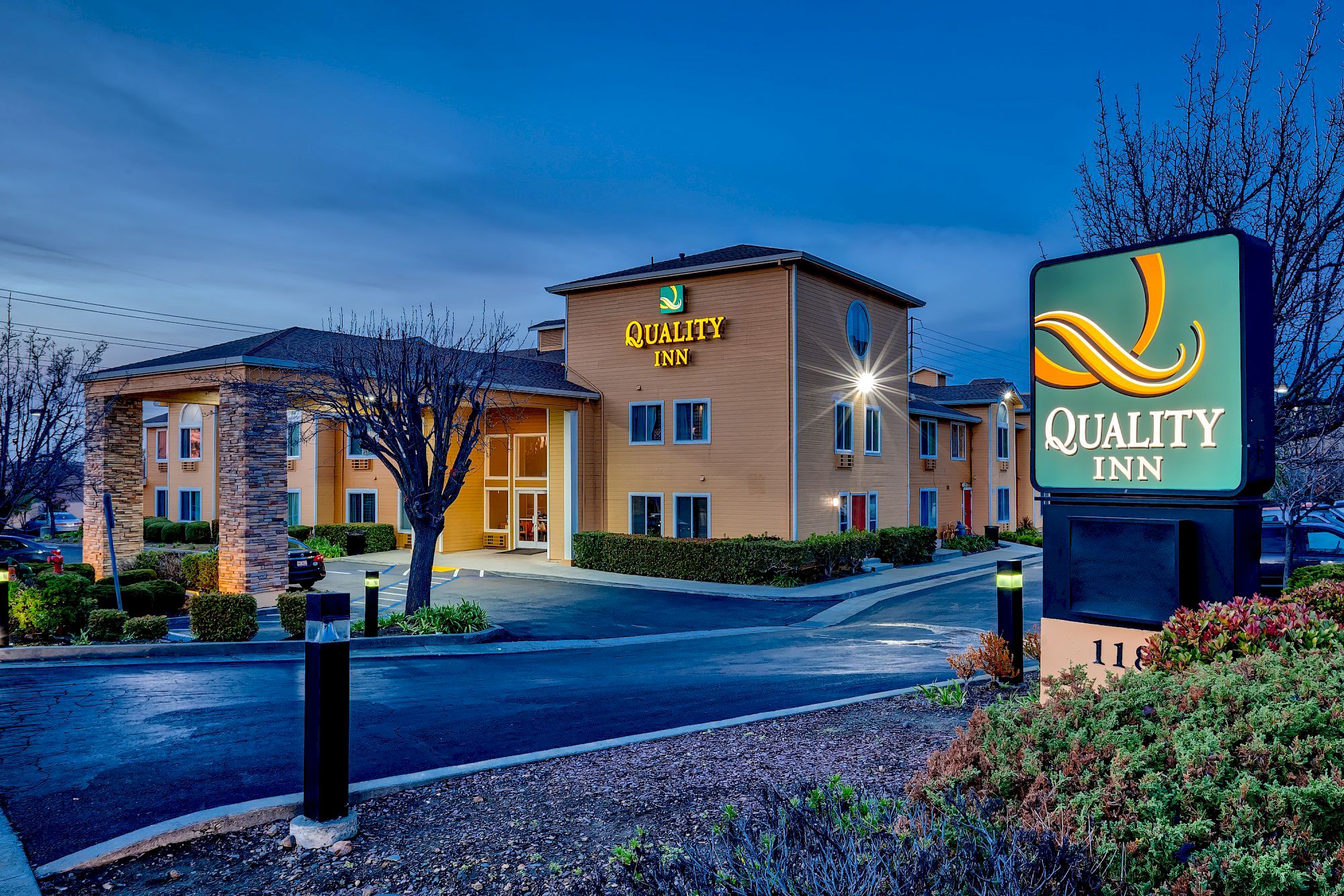 The image shows an exterior view of a Quality Inn hotel at dusk, with illuminated signs and a landscaped entrance area.