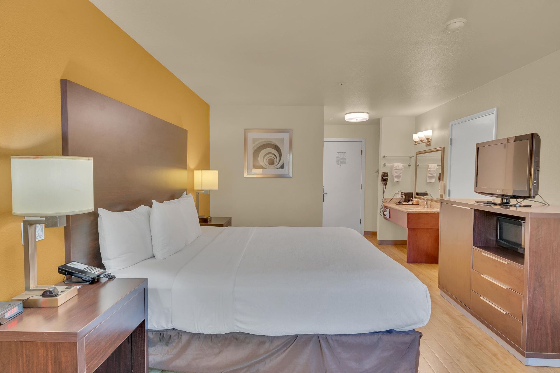A hotel room with a large bed, side tables, lamps, a TV, and a small kitchen area. Walls are light-colored with a decorative picture.