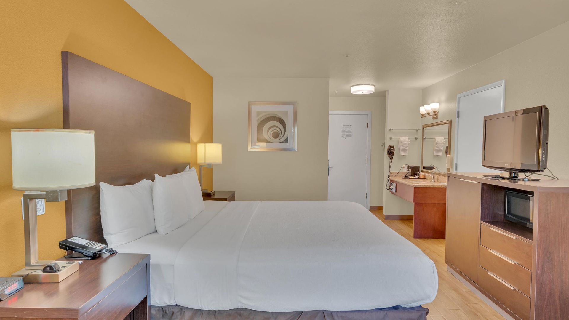 A hotel room with a large bed, bedside tables, lamps, a TV on a cabinet, and a bathroom area with a sink and mirror in the background.