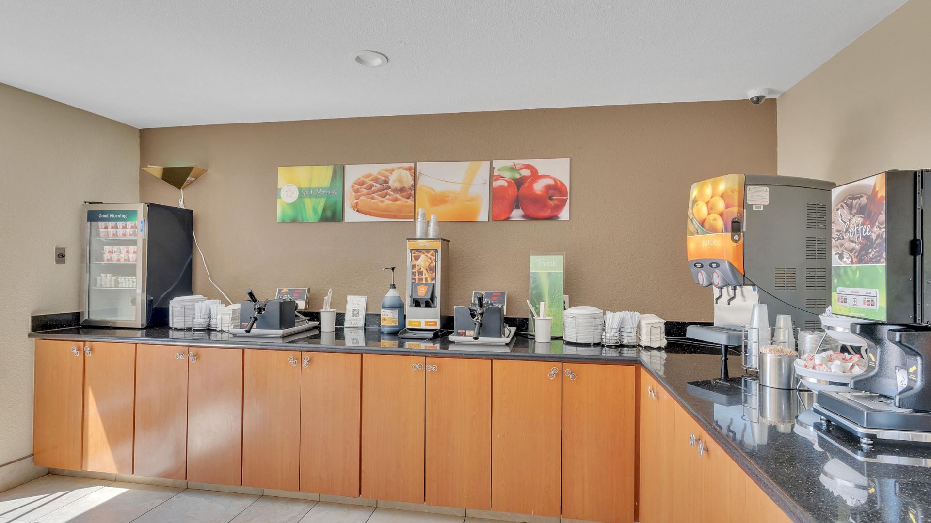 The image shows a breakfast bar setup with beverages, dispensers, and condiments on a countertop in a well-lit room.