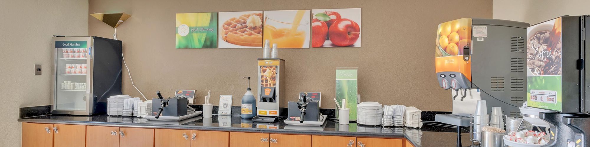The image shows a breakfast bar setup with beverages, dispensers, and condiments on a countertop in a well-lit room.