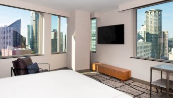 A modern hotel room features a bed, armchair, wall-mounted TV, and city views through large windows ending the sentence.