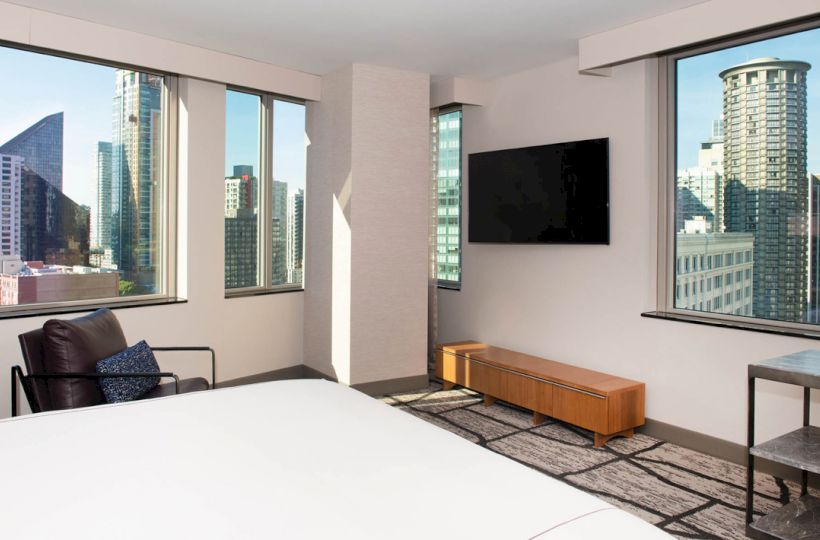 A modern hotel room features a bed, armchair, wall-mounted TV, and city views through large windows ending the sentence.
