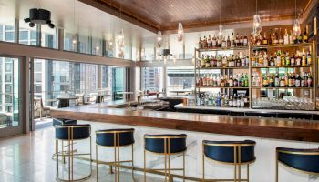 The image shows a modern bar with a large selection of bottles on display, elegant seating, and a view of the city through large windows.