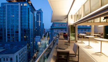 The image shows a modern cityscape at dusk, with a view from a building's balcony featuring outdoor seating and adjacent tall buildings illuminated.