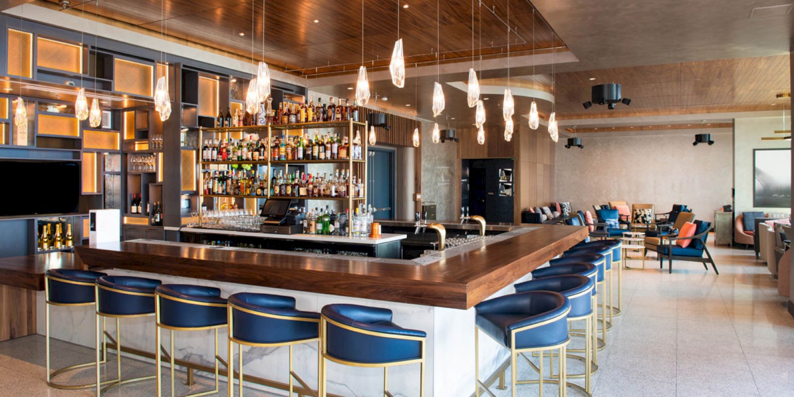 This image shows a modern bar with blue barstools, hanging lights, and a well-stocked liquor shelf. The space has additional seating areas.