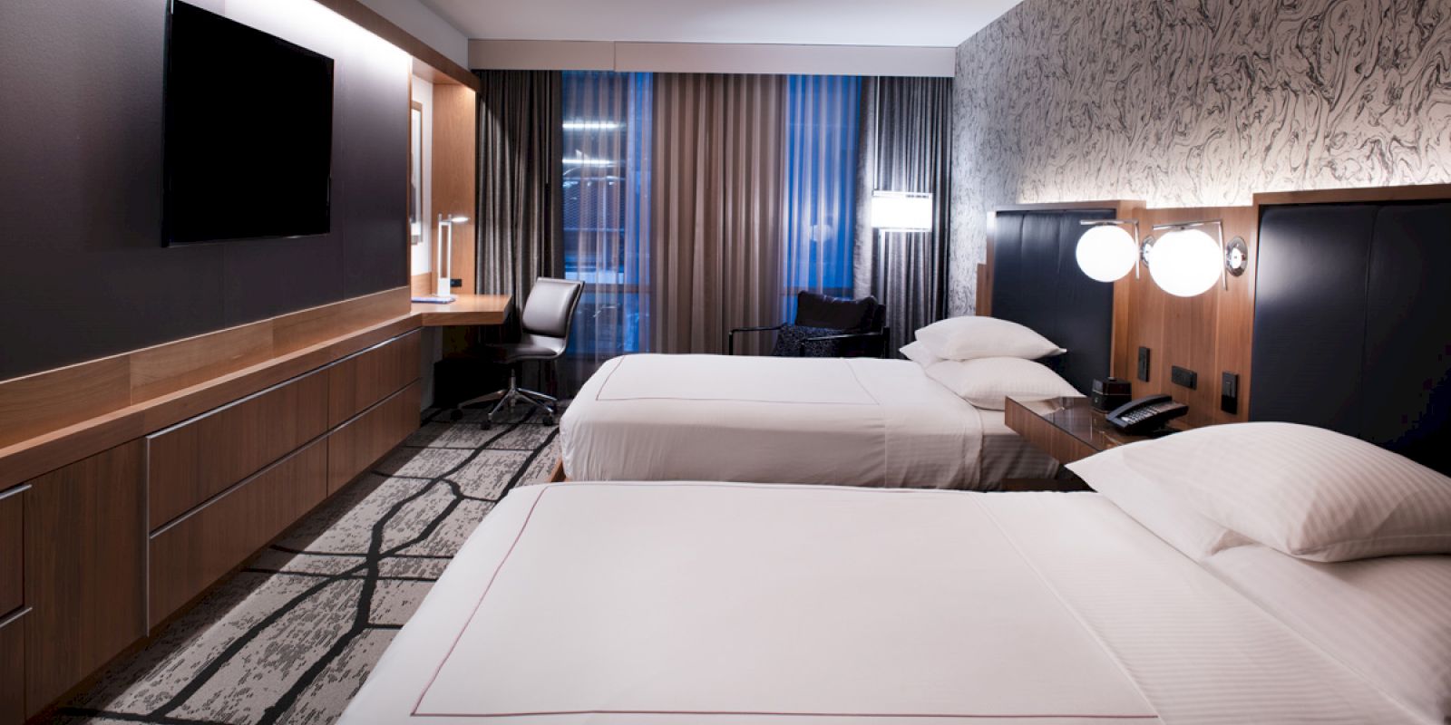 The image shows a modern hotel room with two beds, a wall-mounted TV, a desk with a chair, lamps, and contemporary decor, all neatly arranged.