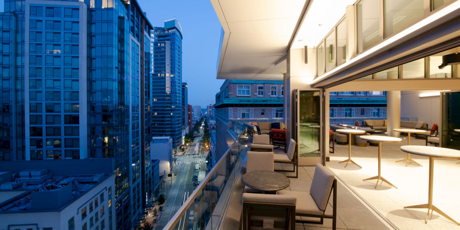 A modern cityscape at dusk with tall buildings, seen from a rooftop patio that has seating areas and tables along the edge.