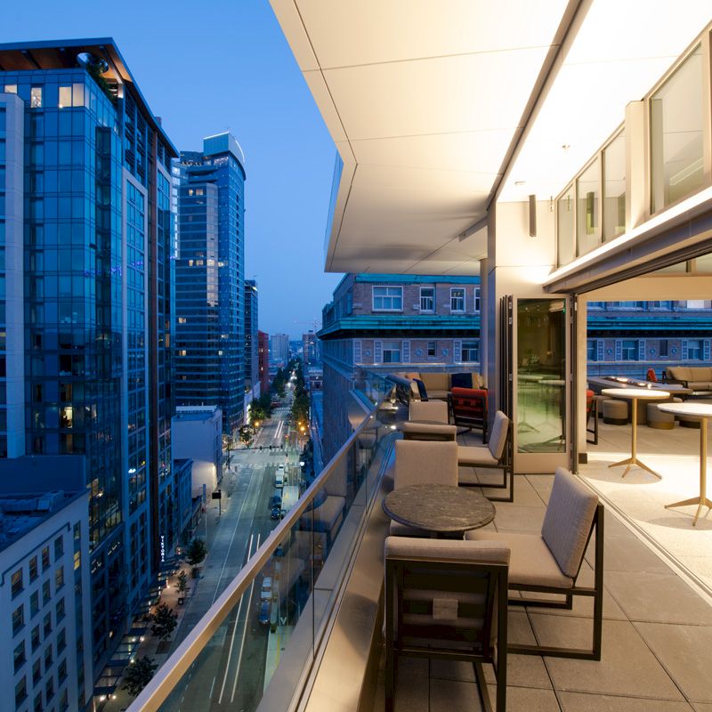 The image depicts a modern cityscape at dusk, with a view from a spacious balcony featuring outdoor seating and tables overlooking tall buildings.