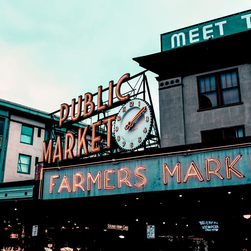 The image shows the iconic neon signs of a public market and a farmers market, along with the facade of surrounding buildings and a partly visible clock.